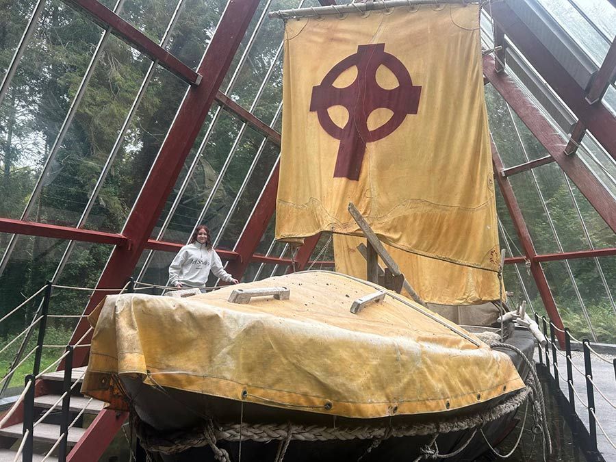 Exposición de un barco recubierto de oro con una gran vela en forma de cruz celta en un espacio museístico con arco de cristal.