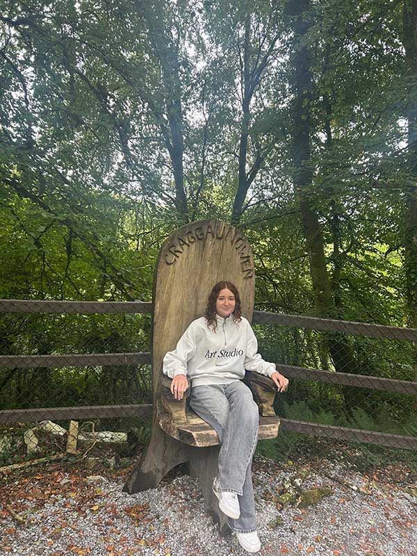 Persona sentada en una silla hecha con un gran tocón de árbol en un bosque frondoso, vestida con un atuendo gris claro y zapatillas blancas.