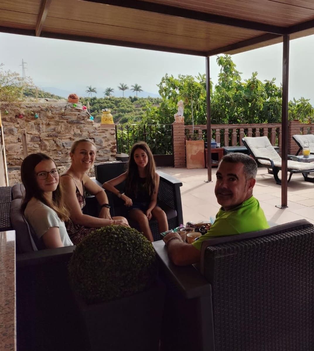 Cuatro personas sentadas en muebles de exterior bajo una pérgola, con vistas a una ladera.