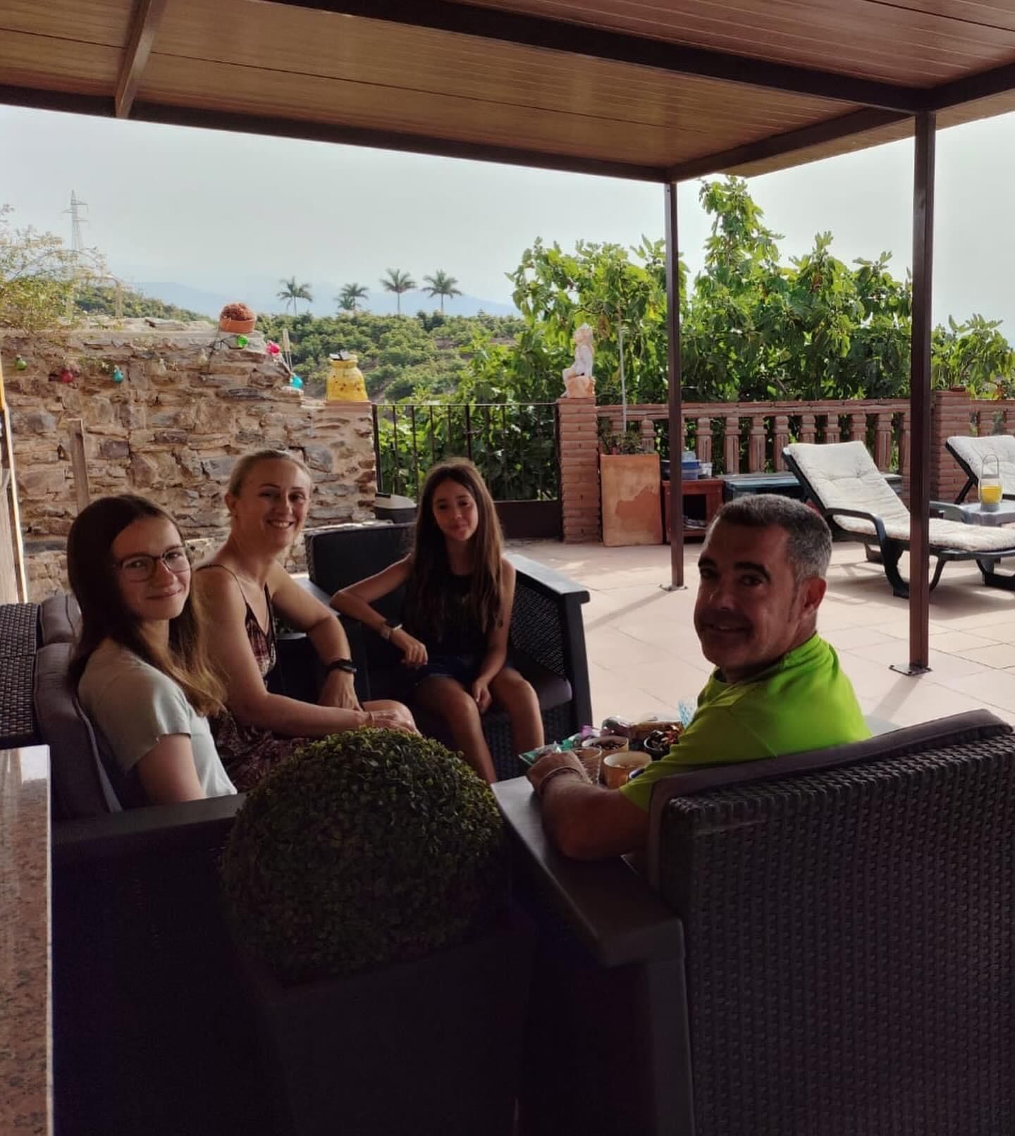 Cuatro personas sentadas en muebles de exterior; un patio soleado con vista a la montaña.