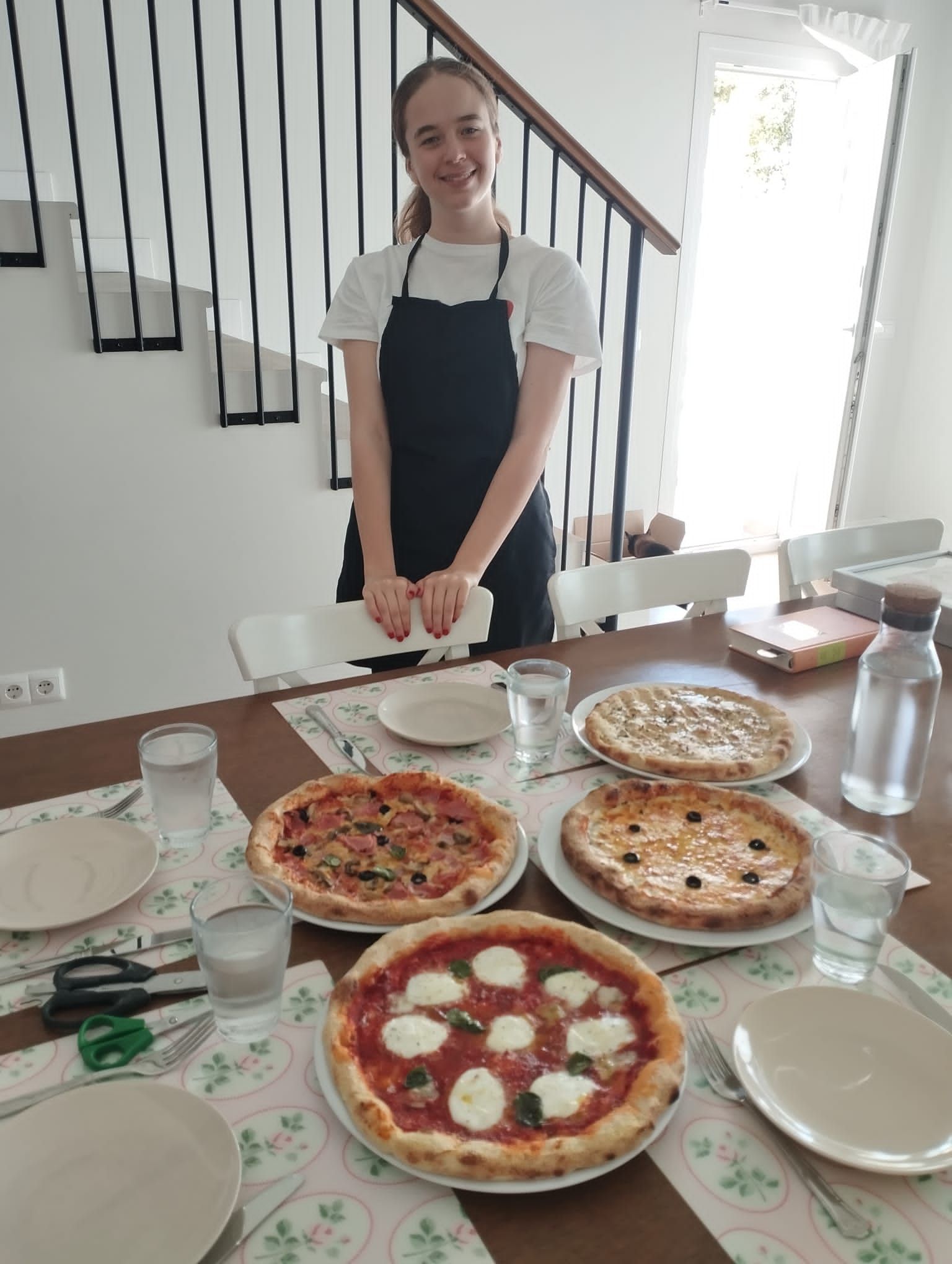 Mujer con pizzas en una mesa, vasos de agua, platos y una escalera al fondo.