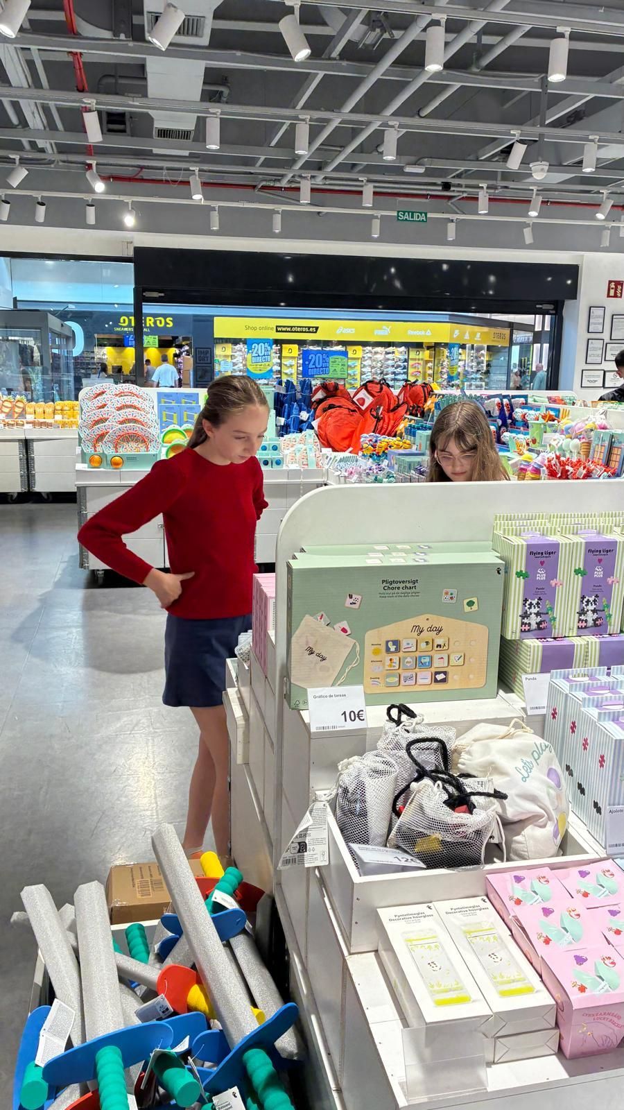 Dos chicas observan artículos expuestos en una tienda bien iluminada. Una viste de rojo, la otra tiene el pelo rizado.