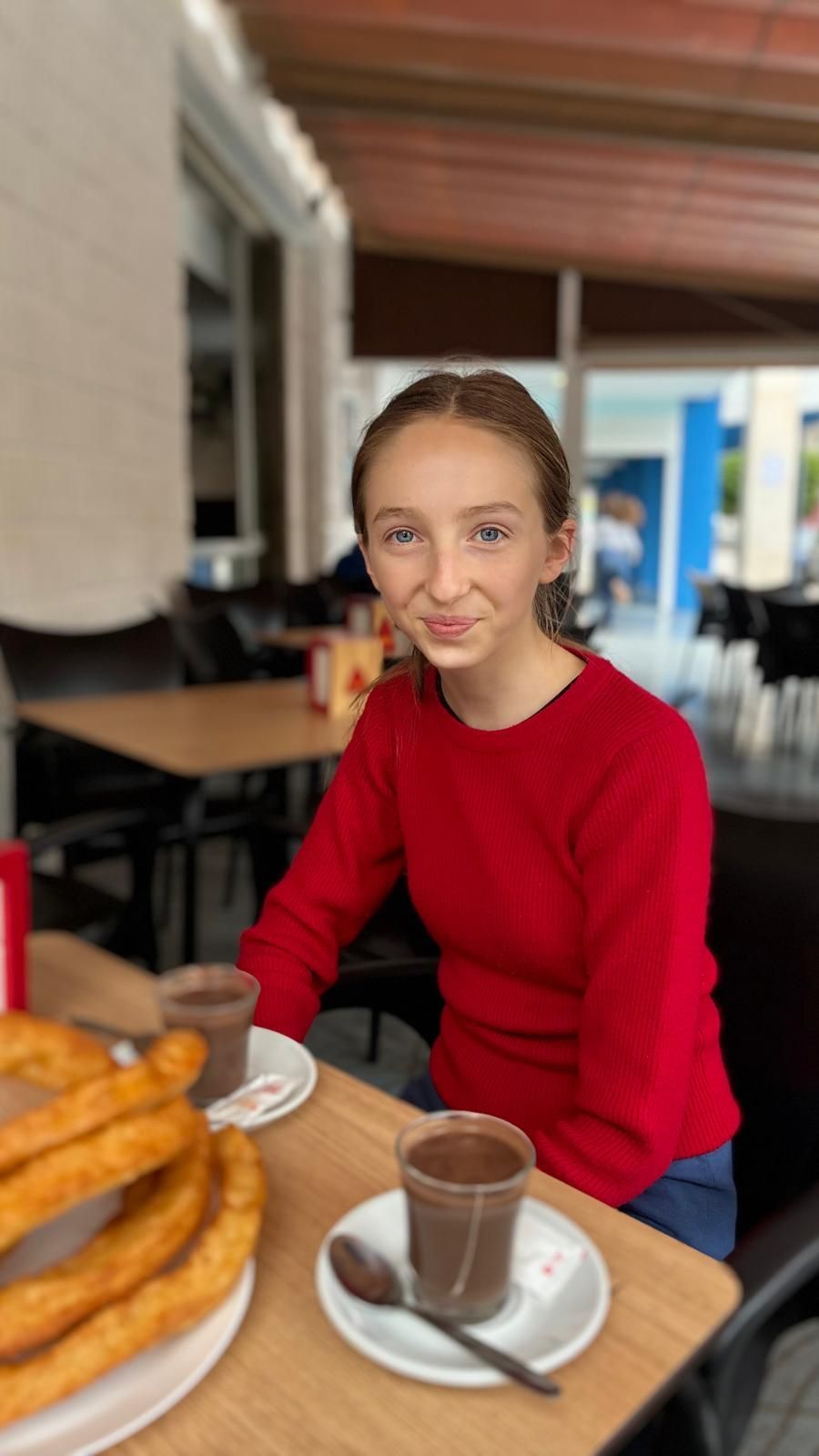 Mujer con suéter rojo sentada en una mesa con churros y chocolate caliente en un café al aire libre.