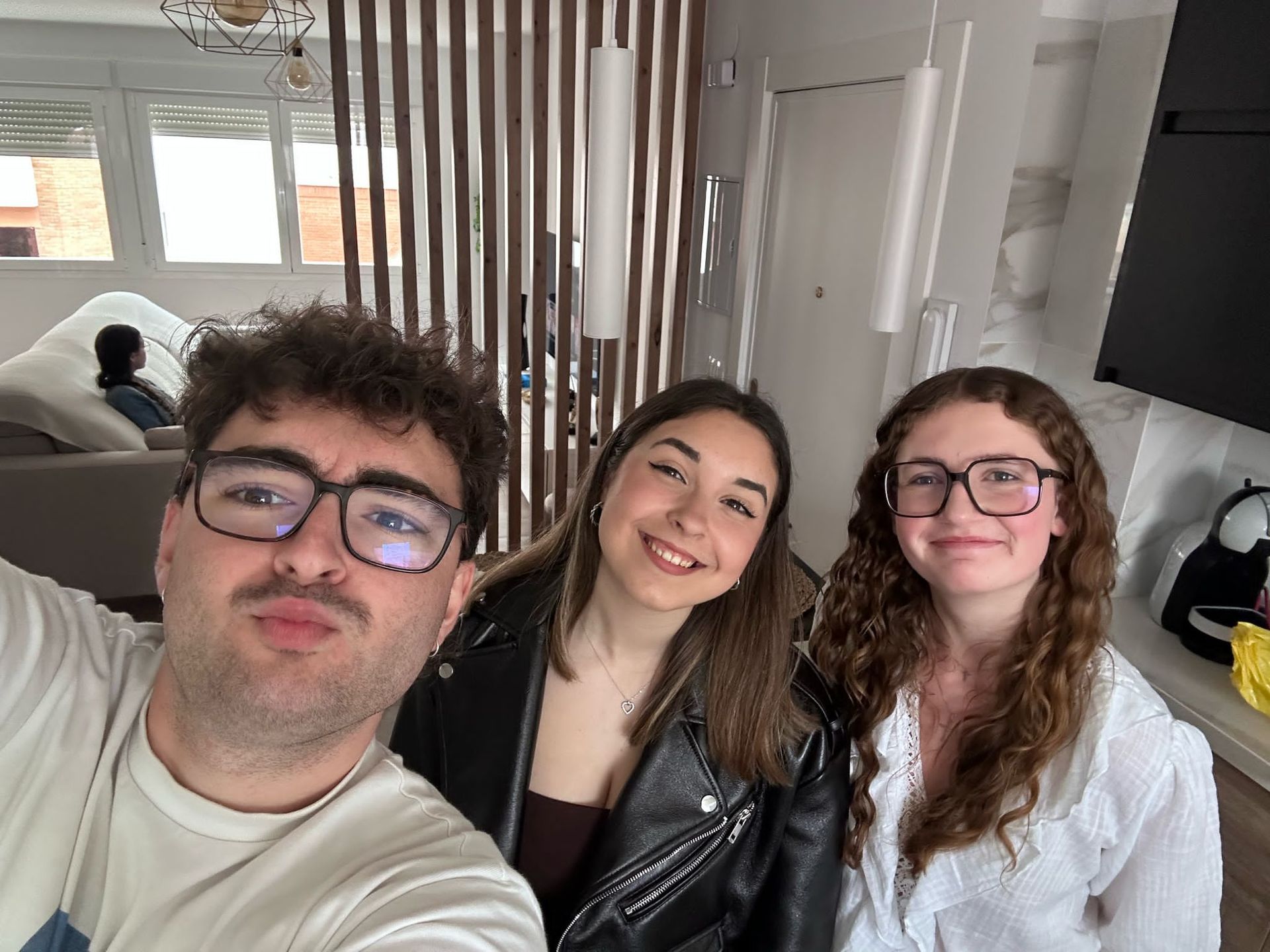 Tres jóvenes adultos sonriendo en un espacio cerrado, tomándose una selfi. Uno lleva gafas y camisa blanca, otro una chaqueta negra y el tercero una bata blanca.