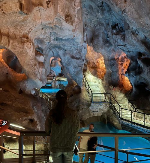 Interior de la cueva con formaciones rocosas iluminadas, barandillas y gente observando.