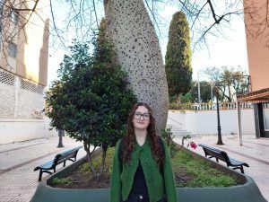 Una mujer con una chaqueta verde está parada frente a un gran árbol con un tronco texturizado, bancos y un edificio de color claro en el fondo.