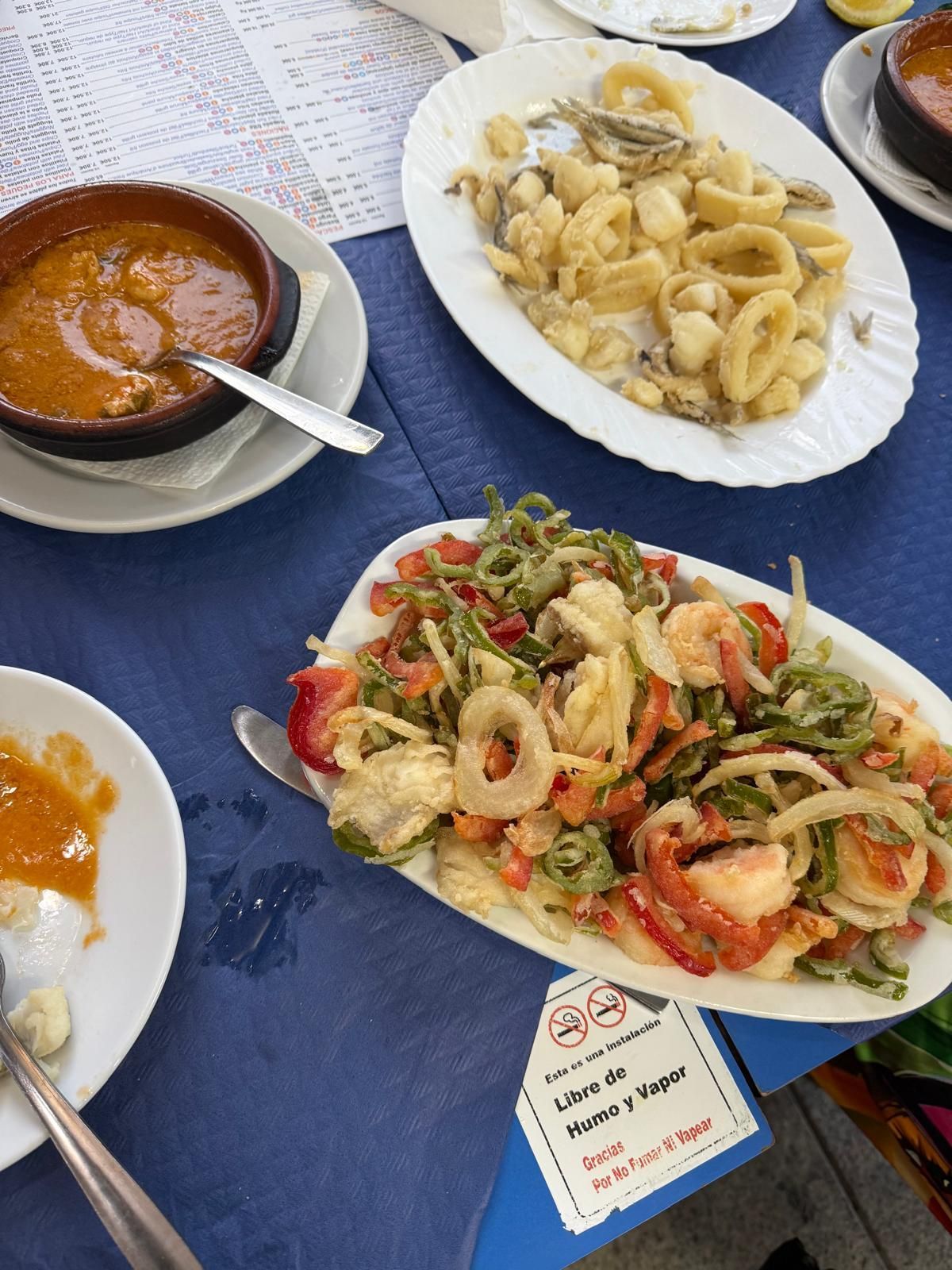 Platos de mariscos sobre mantel azul: calamares fritos, ensalada de camarones y verduras y sopa.