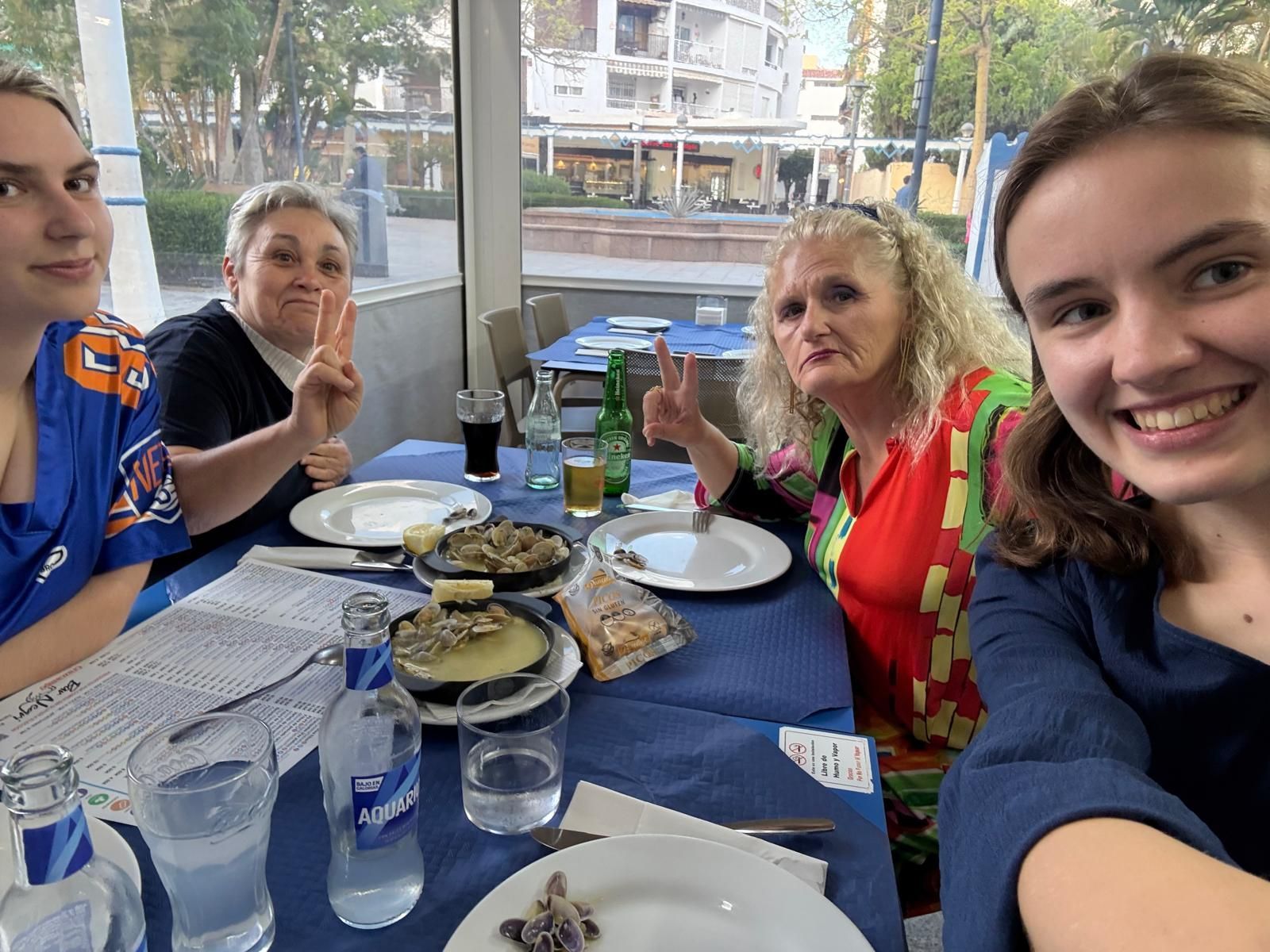 Cuatro personas sentadas a una mesa con comida, bebidas y un mantel azul. Sonríen y gesticulan.