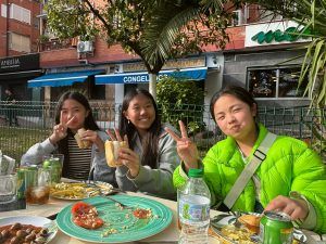 Tres personas en una mesa de café al aire libre, sonriendo y sosteniendo el símbolo de la paz. Hay comida y bebida en la mesa.