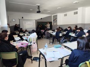 Escena de aula con estudiantes en sus escritorios, un profesor presentando cerca de una pantalla y un reloj en la pared.