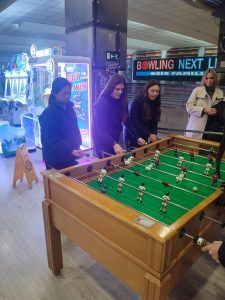 Gente jugando al futbolín en una galería. Cartel de bolos al fondo.