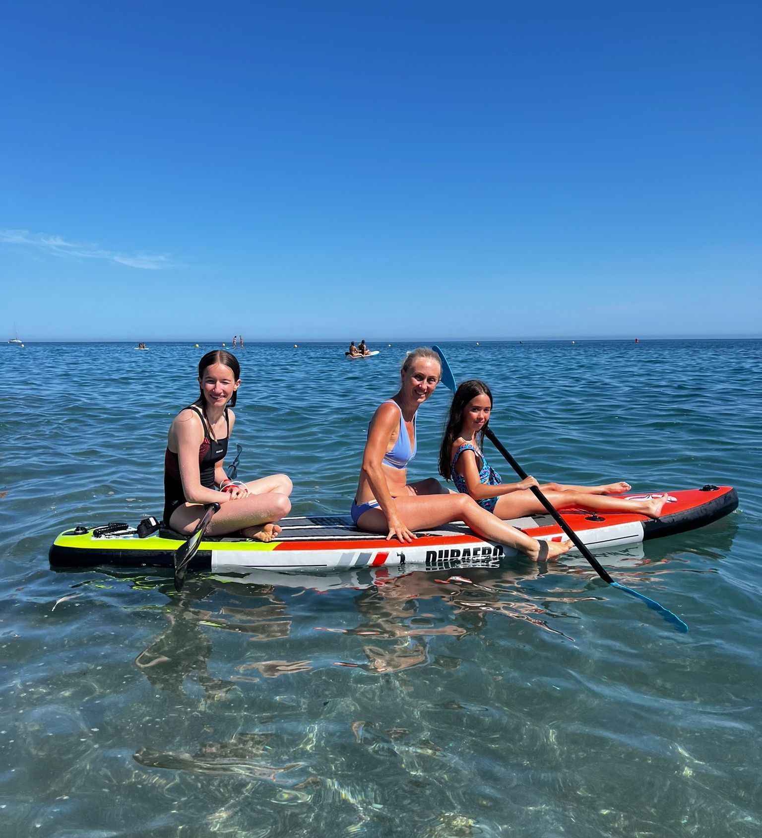 Tres personas en una tabla de paddle surf en aguas cristalinas bajo un cielo azul; están sonriendo.