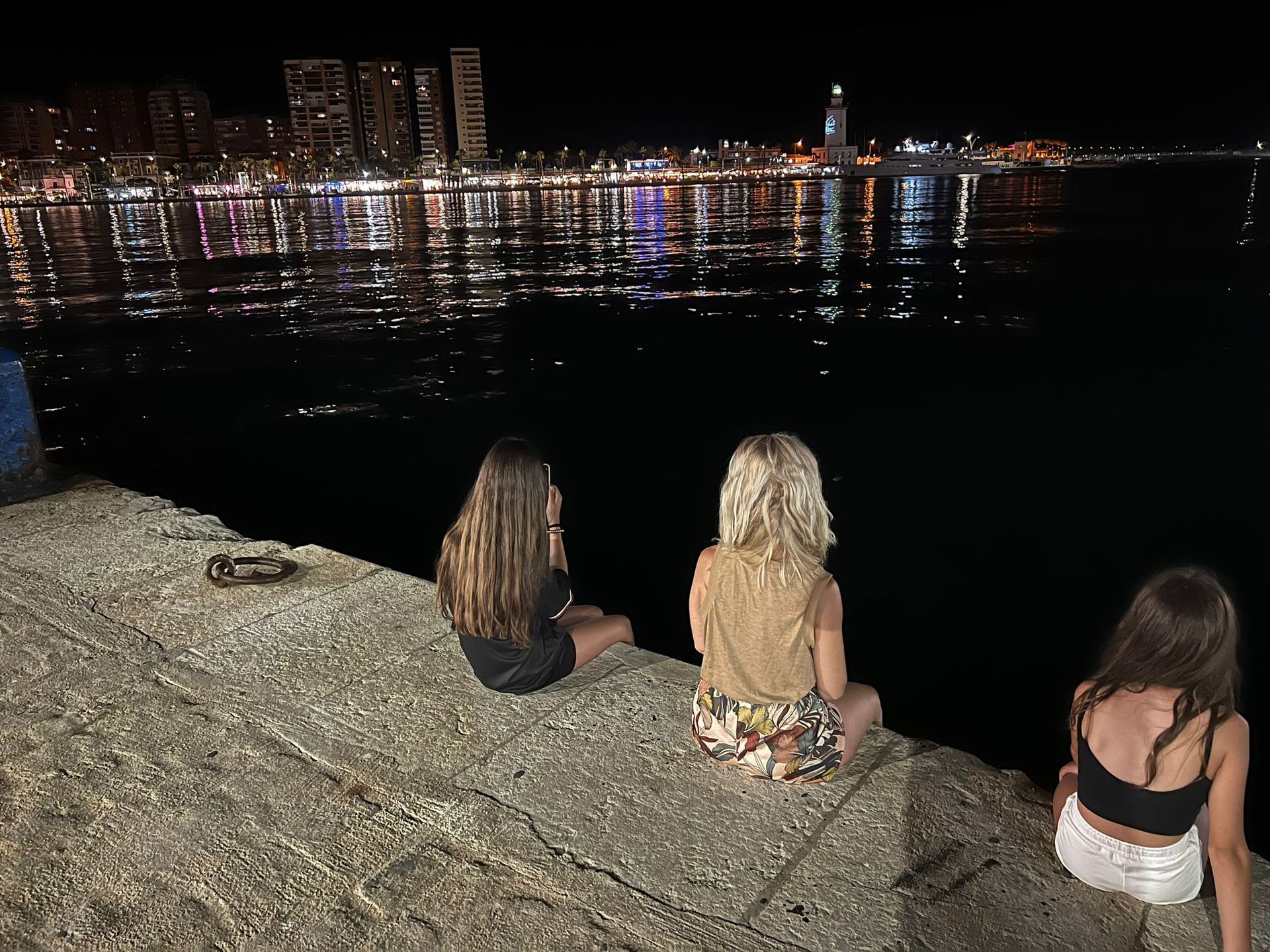 Tres personas se sientan en una cornisa de hormigón, mirando el horizonte de una ciudad nocturna reflejada en el agua.