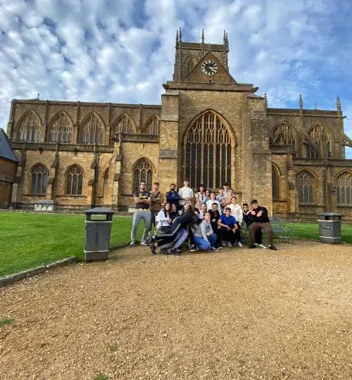 Un grupo de personas posa frente a una gran iglesia de piedra. Cielo nublado. Camino de grava marrón.