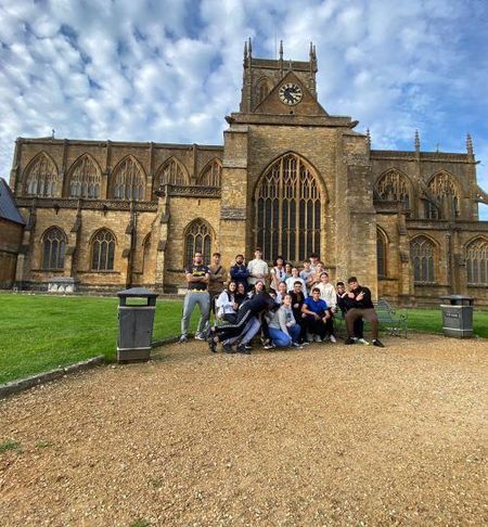 Grupo posando sobre grava frente a una gran iglesia histórica con torre del reloj bajo un cielo azul nublado.