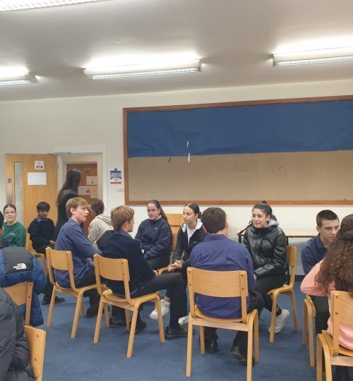 Grupo de estudiantes sentados en sillas, conversando. Aula con alfombra azul y panel mural.