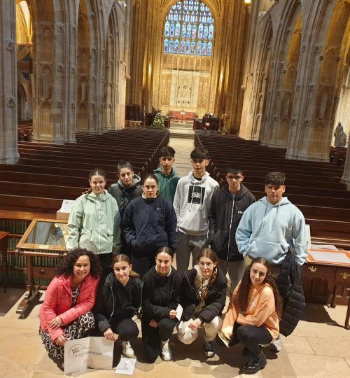 Grupo de adolescentes dentro de una catedral, posando para una foto. Iluminación tenue, bancos y un gran vitral.