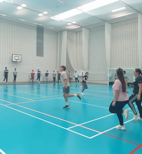 Personas haciendo ejercicio en un gimnasio con suelo azul, paredes blancas y red.