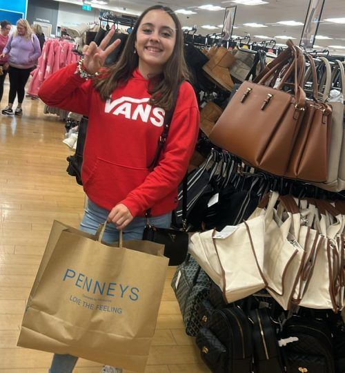 Una mujer con una sudadera con capucha roja de Vans sostiene una bolsa de compras de Pennys, haciendo un signo de paz, en una tienda con bolsos.