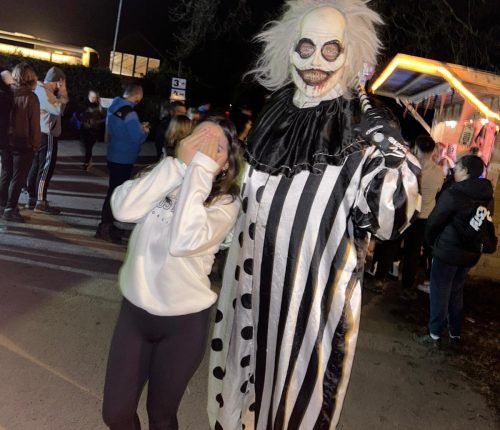 Niña cubriéndose la cara, asustada por un payaso alto con traje de rayas blancas y negras, en un evento nocturno.