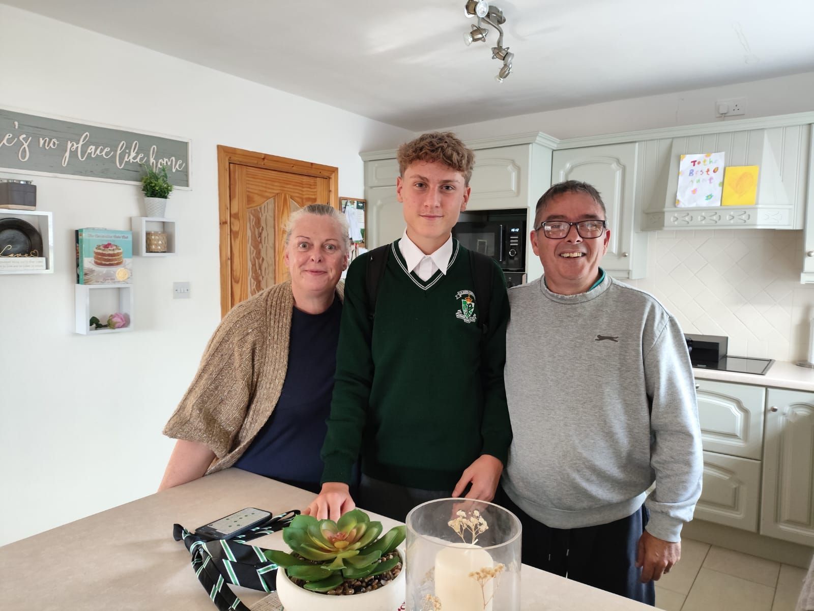 Una familia de tres sonriendo en una cocina, un adolescente con uniforme verde se interpone entre sus padres.