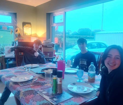 Tres personas sentadas a una mesa preparada para comer. Las ventanas muestran un coche afuera.