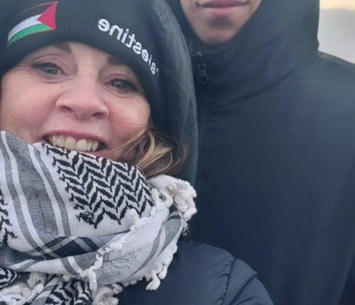 Mujer con sombrero con bandera palestina y bufanda keffiyeh, sonriendo junto a una persona con una chaqueta negra.