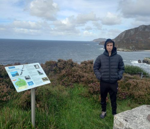 Hombre de pie junto a un cartel con el océano y las colinas al fondo, vestido con una chaqueta negra y las manos en los bolsillos.