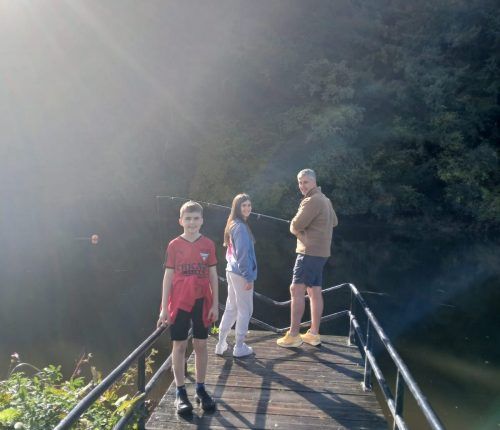 Tres personas están de pie en un muelle de madera sobre el agua. Luz solar brillante.