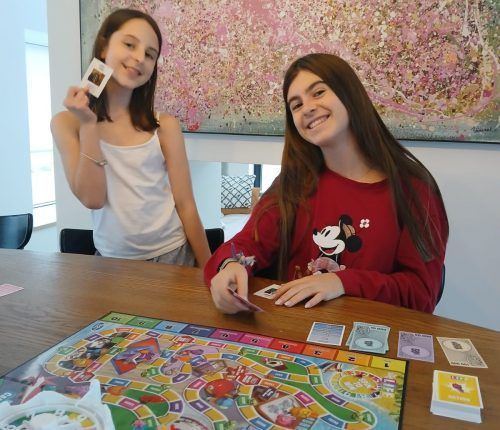 Dos niñas juegan a un juego de mesa en una casa. Una sostiene una carta, la otra sonríe con otra en la mano.