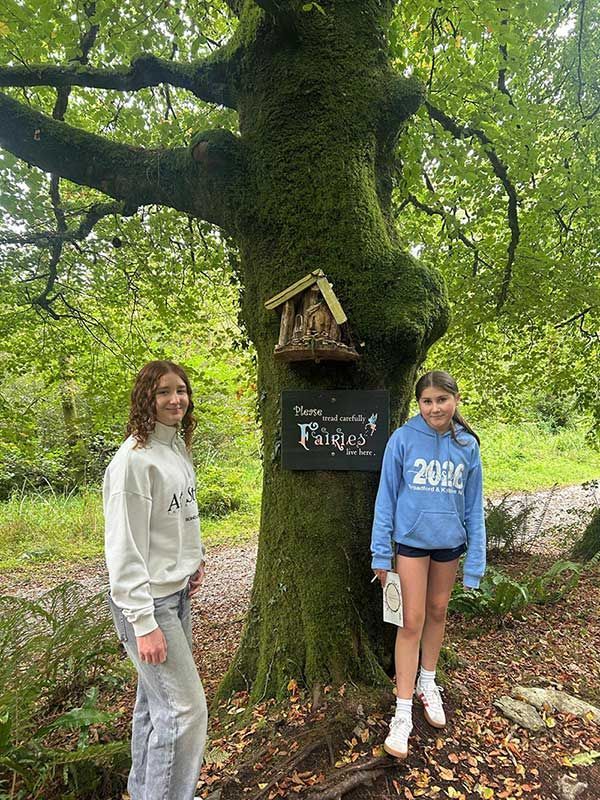 Dos personas de pie junto a un gran árbol con un pequeño cartel y una casita para pájaros en una zona boscosa.