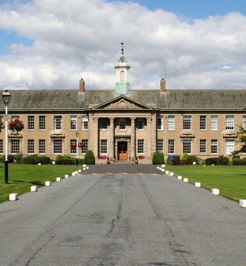 Edificio largo de color beige con columnata, cúpula y entrada. Día soleado, césped verde.