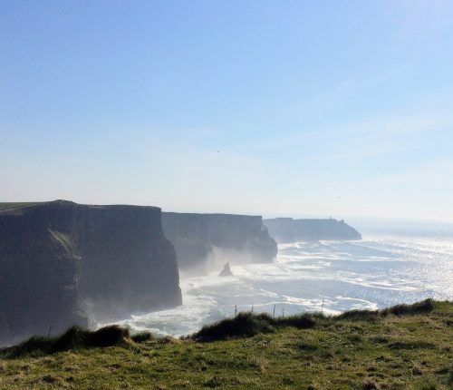 Acantilados de Moher a lo largo de la costa irlandesa con olas rompiendo bajo un cielo azul.