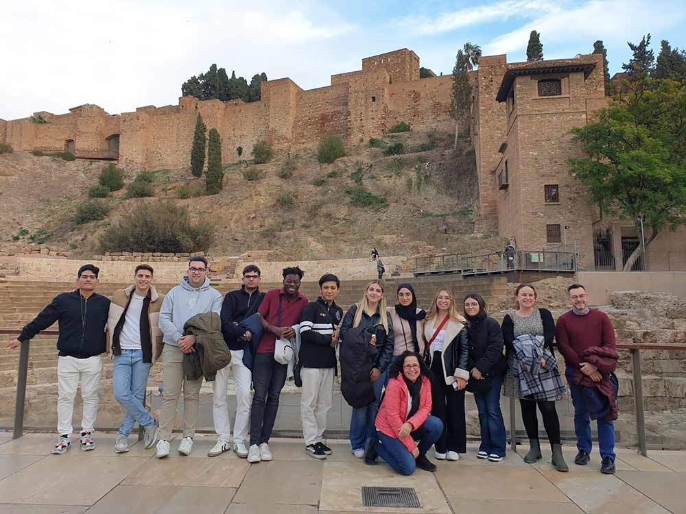 Grupo de personas de pie frente a un edificio de piedra con árboles y una colina al fondo.
