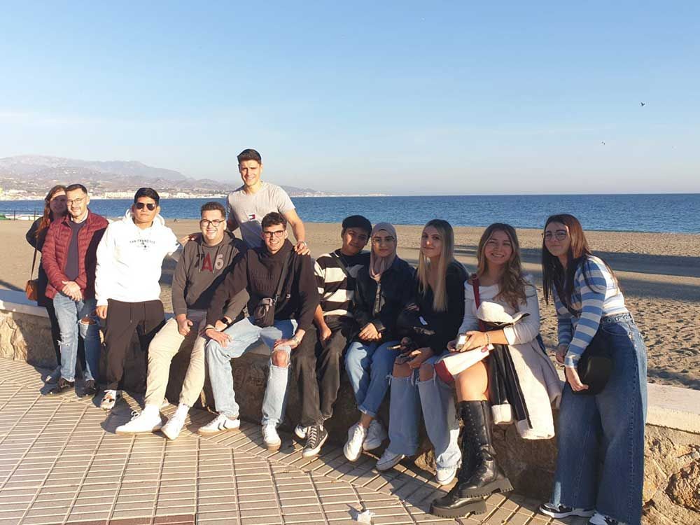 Un grupo de personas posa junto al mar en un día soleado. Están sentadas en un muro, con la playa y las montañas al fondo.