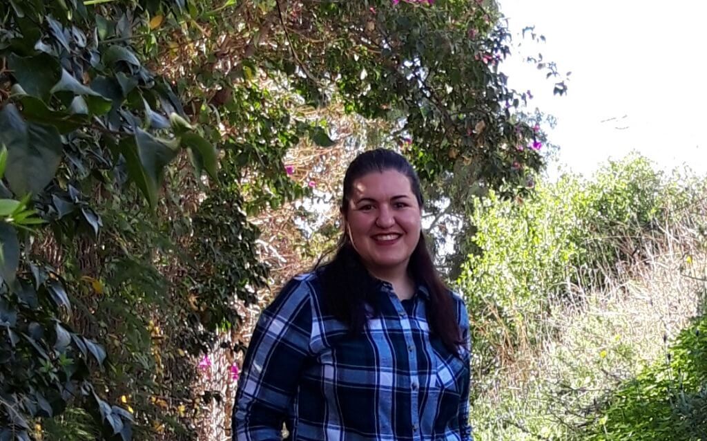 Mujer sonriendo, vistiendo una camisa a cuadros azul, de pie al aire libre con un exuberante follaje verde de fondo.