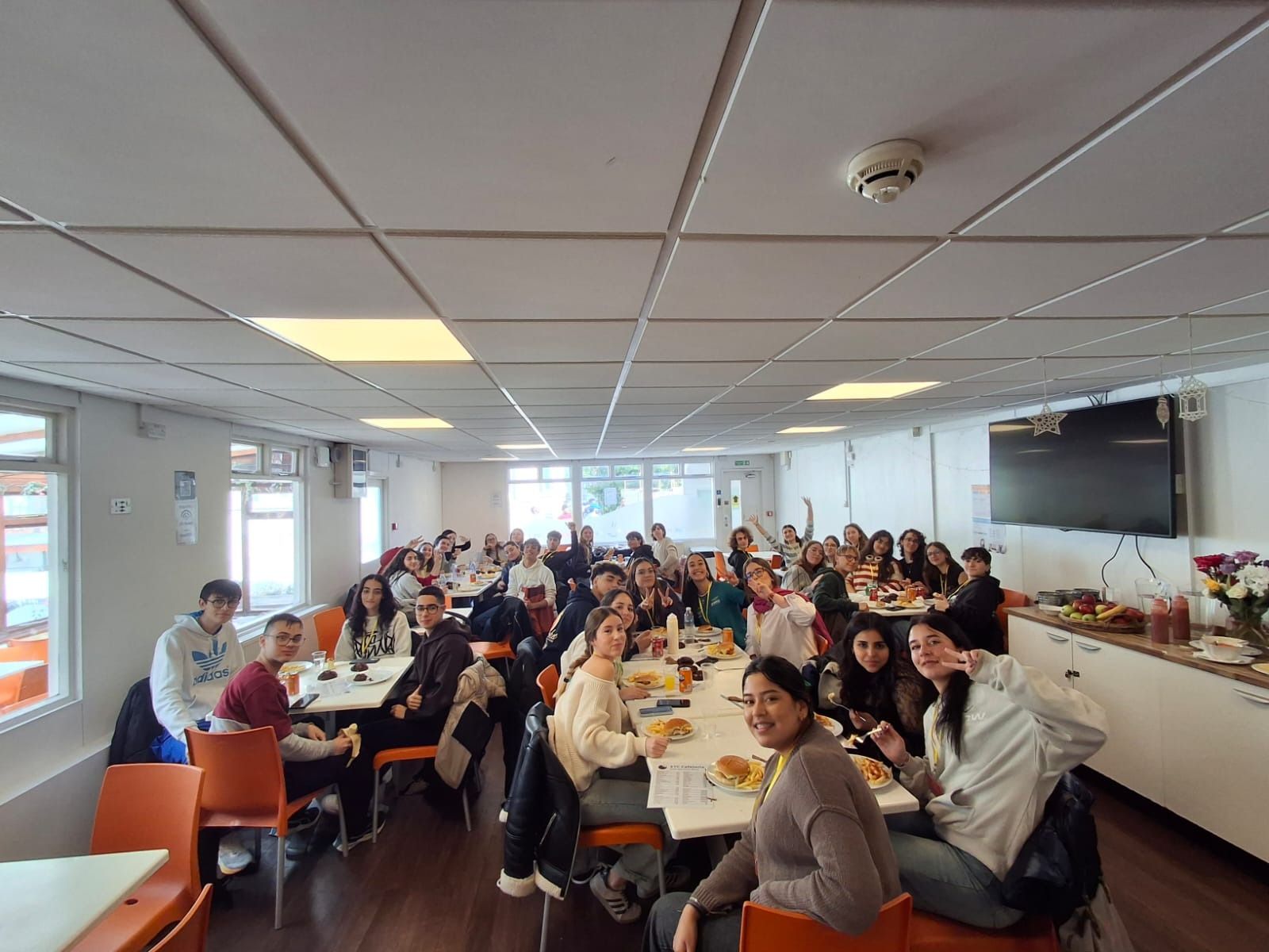 Un grupo de personas se reunió en un comedor, algunas sonriendo y levantando la mano. Espacio luminoso, ventanas y mesas.