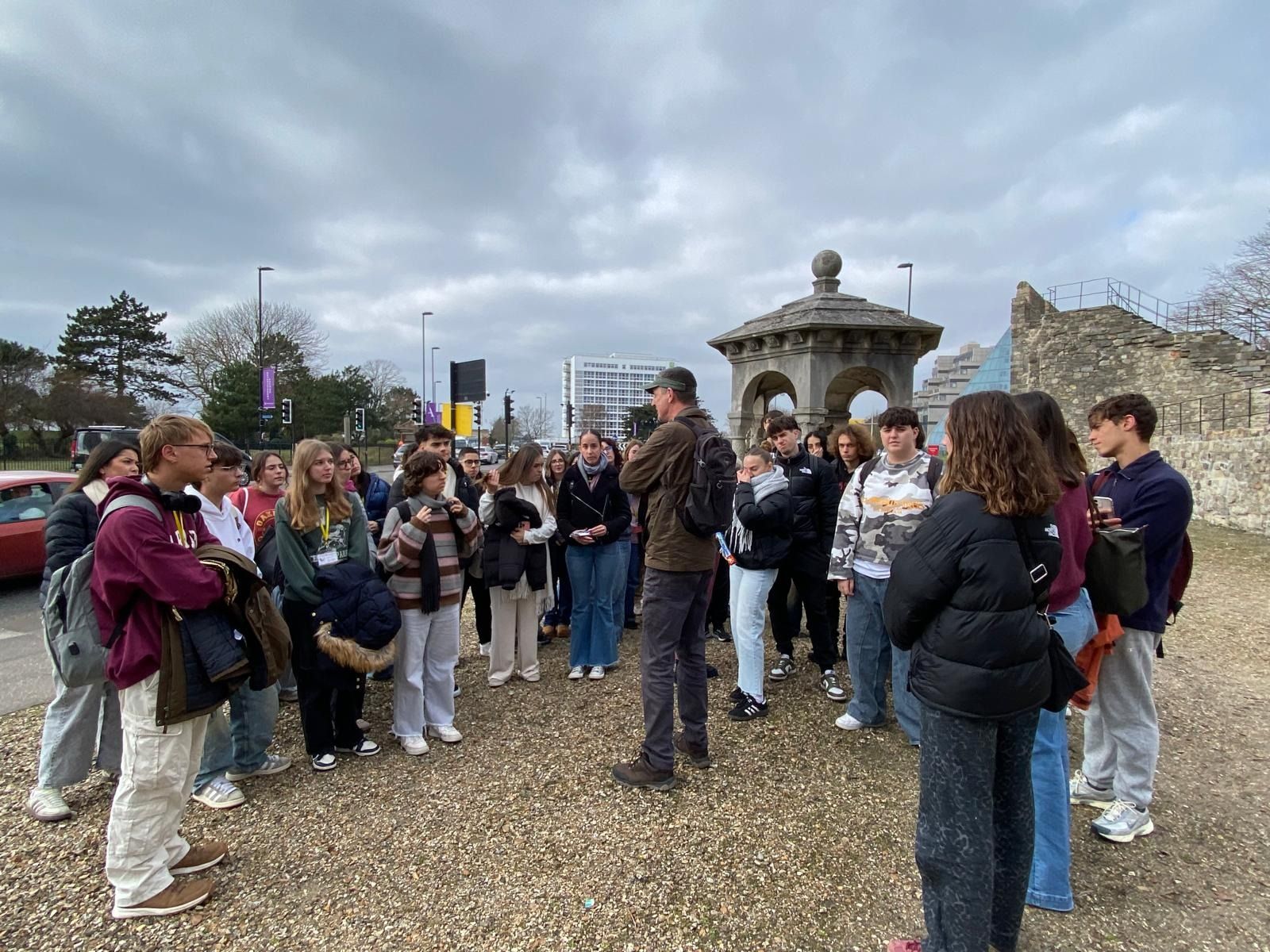 Un grupo de personas escucha a un hombre. Están afuera, junto a una estructura de piedra, en un día nublado.