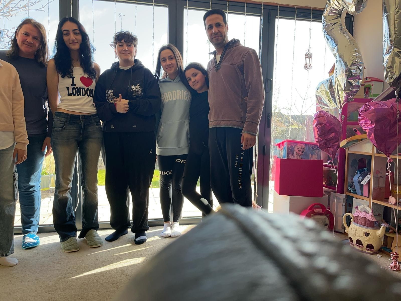 Un grupo de personas sonriendo, posando frente a una ventana. Al fondo, una habitación decorada y globos.