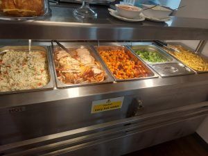 Un buffet con cinco bandejas de comida de metal que contienen diversos platos, detrás de una vitrina de cristal.