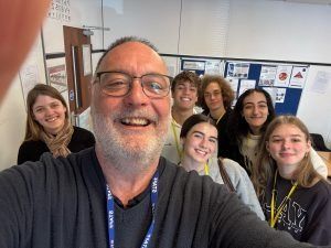 Un hombre se toma un selfie con siete estudiantes sonrientes en un salón de clases.