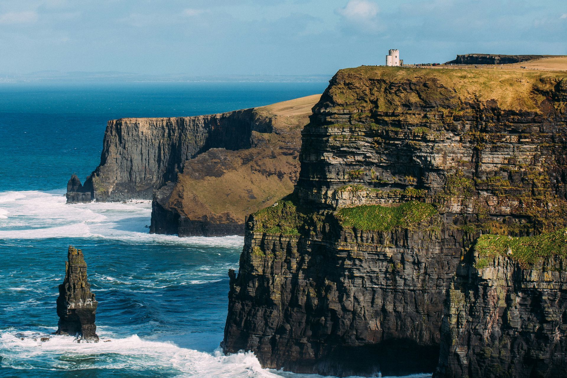 Acantilados de Moher, Irlanda, en contacto con el océano Atlántico. Acantilados escarpados de color verde-marrón con una torre blanca.