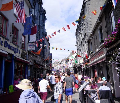 Calle peatonal concurrida con banderas, restaurantes al aire libre y gente caminando.