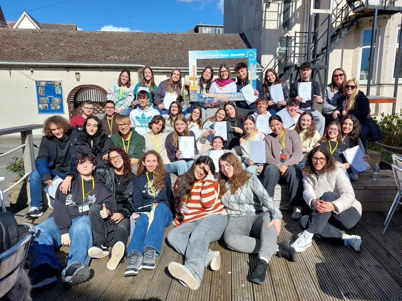 Grupo de jóvenes sosteniendo papeles, sonriendo, posando al aire libre en una plataforma de madera.