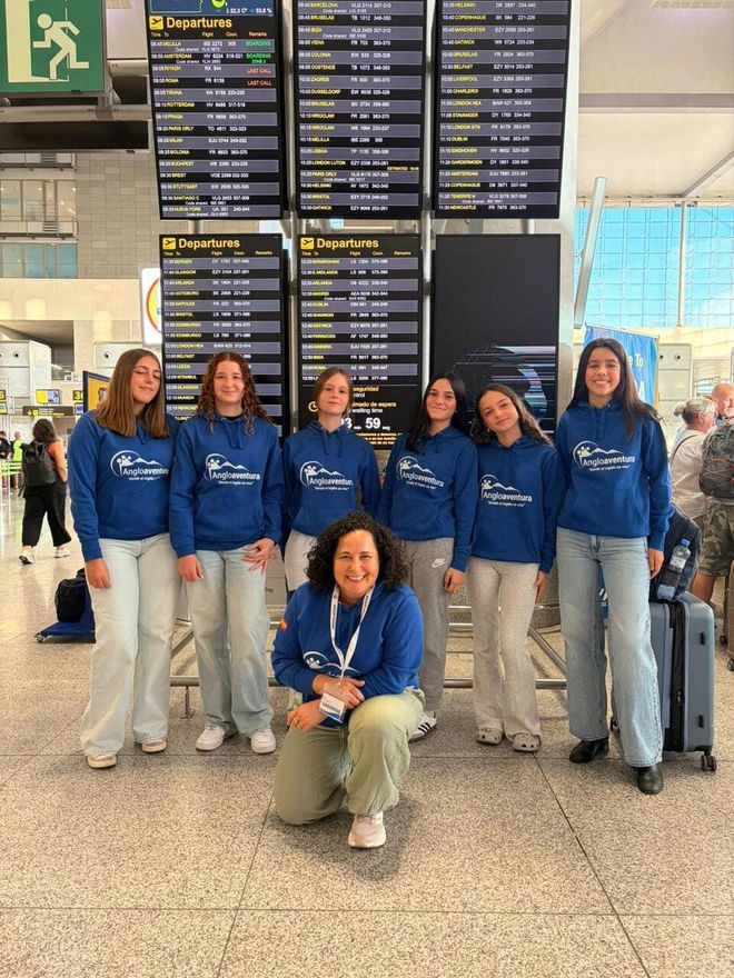 Grupo de personas con sudaderas azules posando en una terminal de aeropuerto bajo los paneles de salidas.