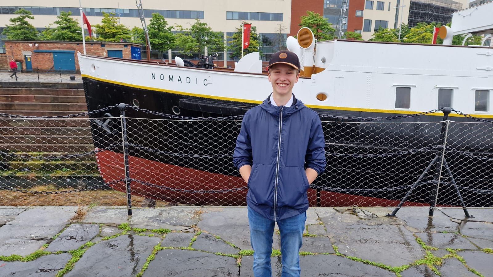 Un joven con chaqueta azul sonríe frente a un barco llamado