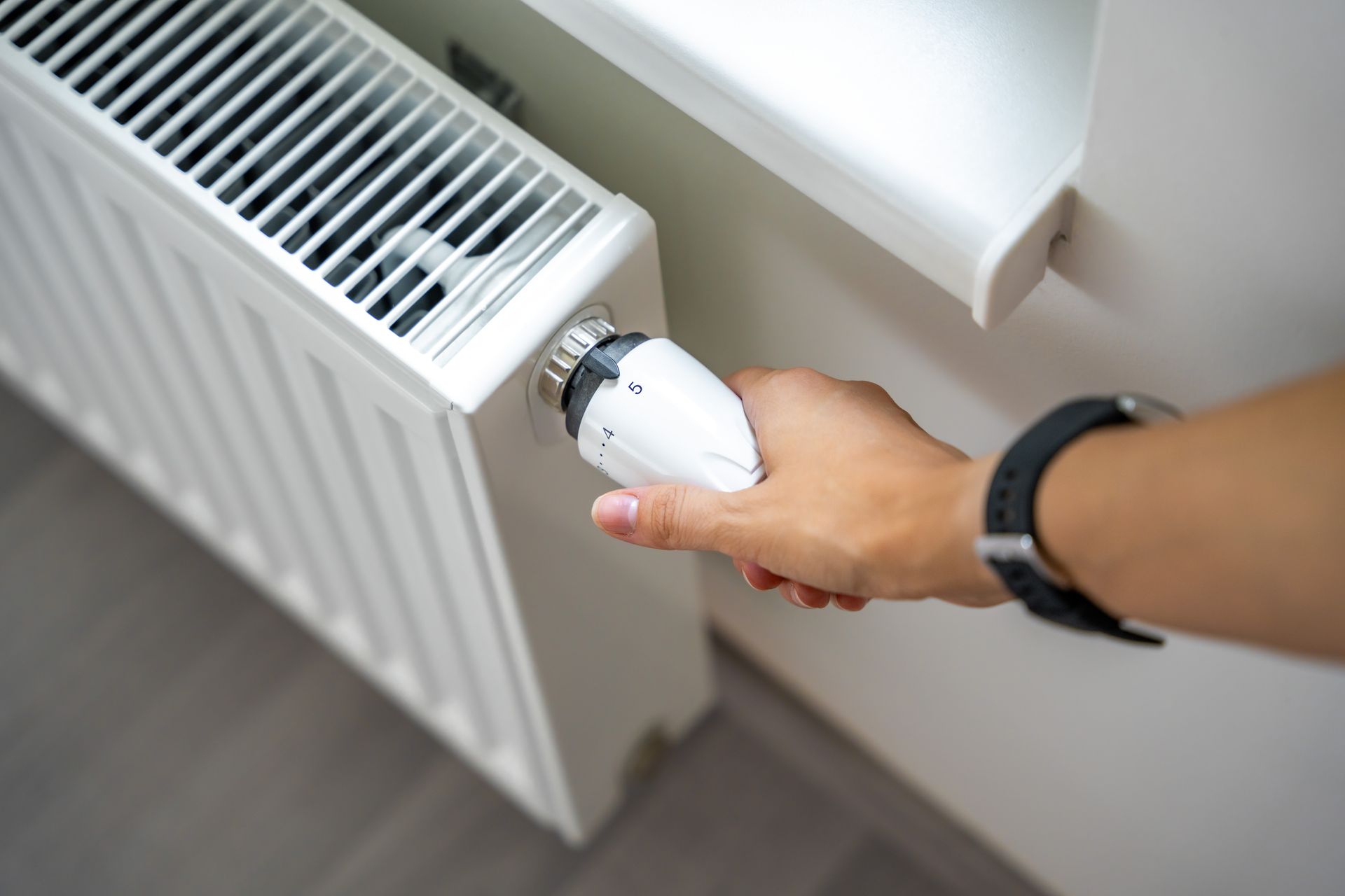 Une main actionne un robinet de radiateur blanc, près d'une fenêtre.