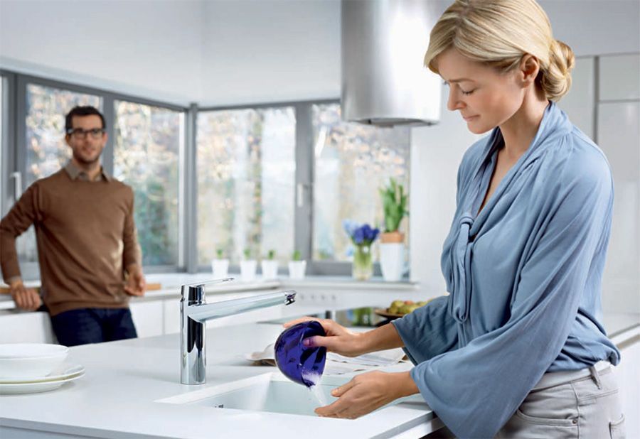 Une femme lave un bol dans un évier de cuisine ; un homme la regarde en arrière-plan.