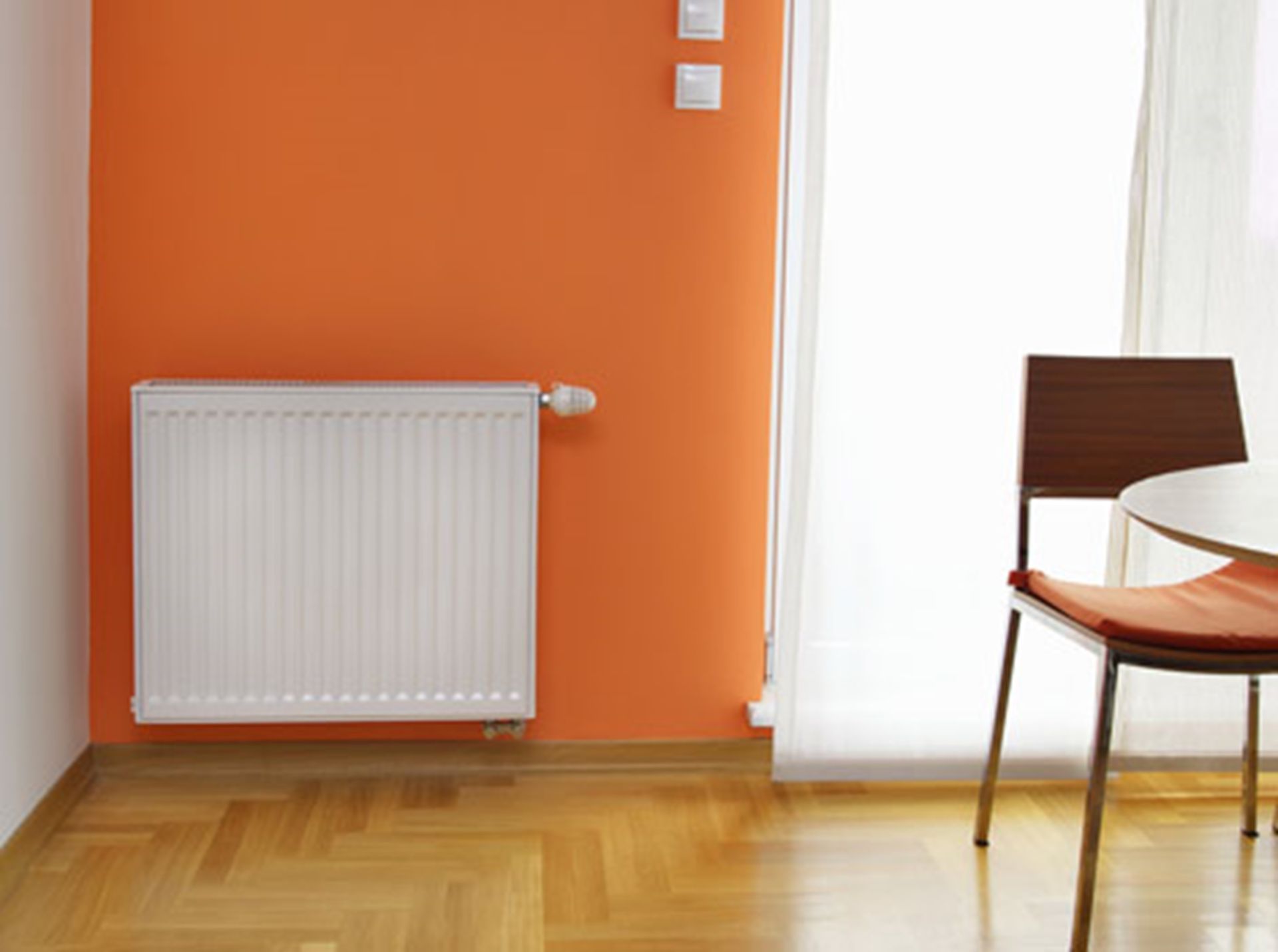 Radiateur blanc contre un mur orange ; chaise près de la fenêtre avec des voilages. Sol en bois.