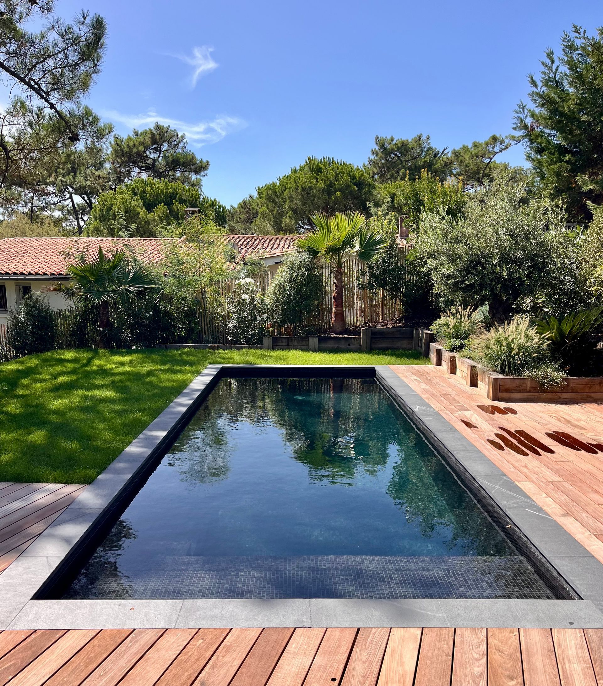 Piscine rectangulaire aux eaux sombres, entour&eacute;e d'une terrasse en bois et d'une pelouse verte, avec des arbres en arri&egrave;re-plan sous un ciel bleu